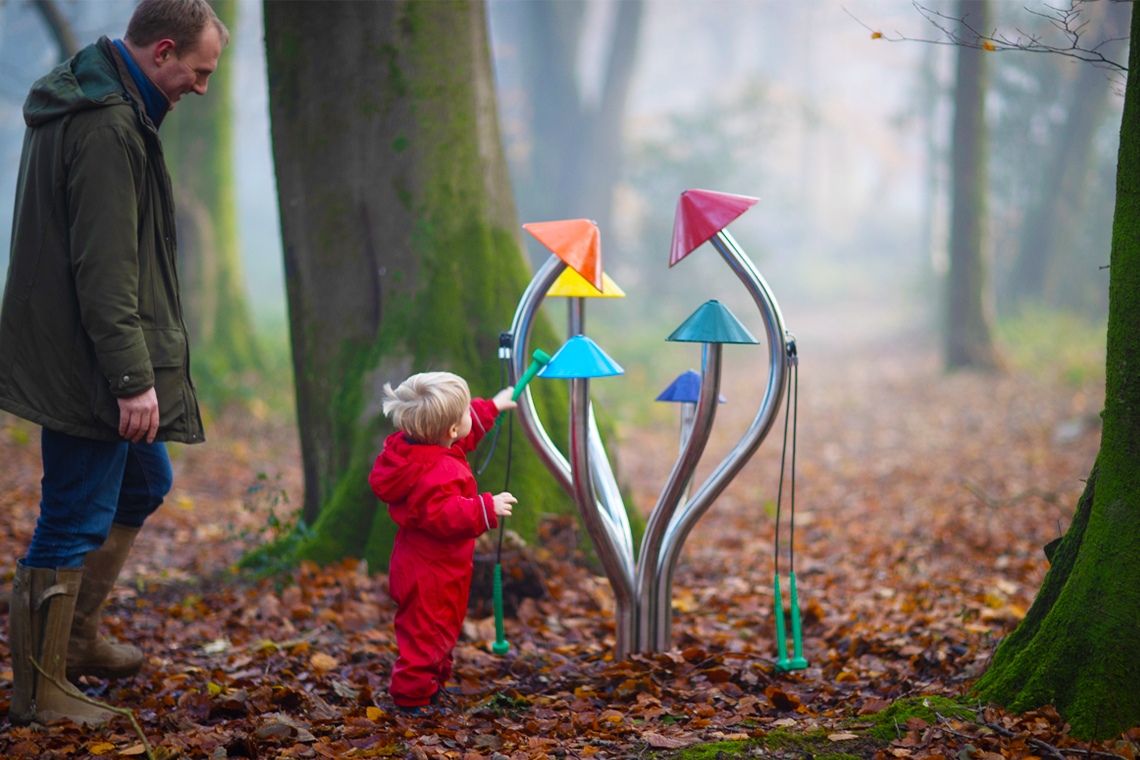 father holding his son who is wearing a red rainsuit and playing outdoor musical chimes shaped like a cluster of colorful mushrooms