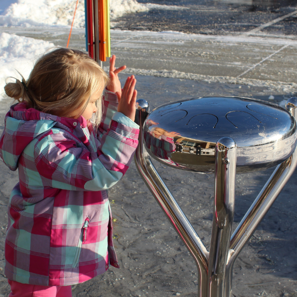 a young girl in a thick coat playing a stainless steel tongue drum outside a the clinton macomb public library