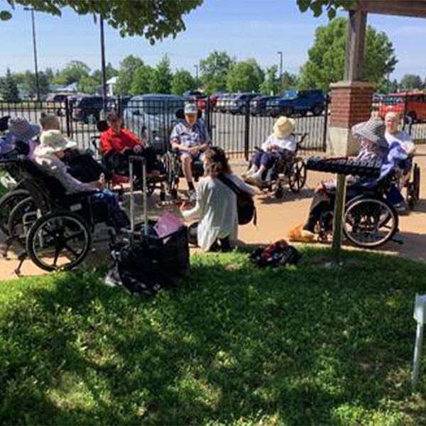 a group of seniors some in wheelchairs gathered around outdoor musical instruments with a music therpist playing guitar