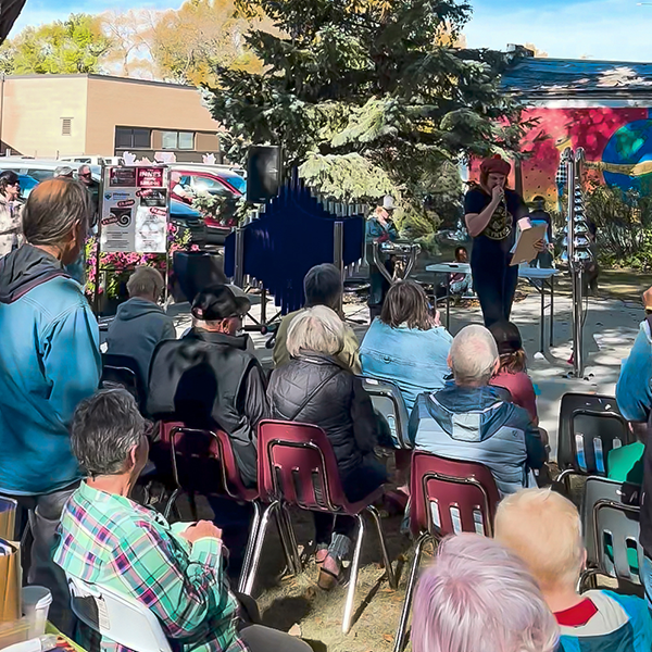 Outdoor community gathering at the opening of a musical playground with a diverse crowd seated, facing a presenter on stage. A vibrant mural and trees in the background set a lively tone.