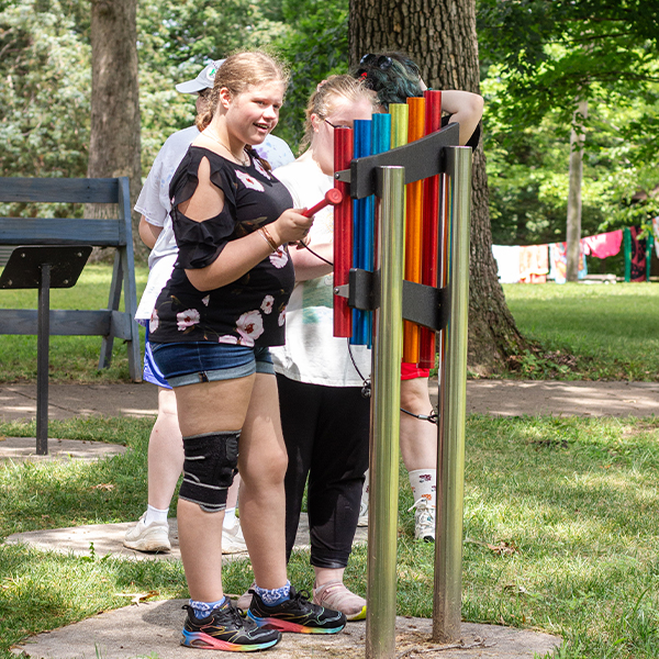 teenage girls with additional needs playing outdoor musical chimes in rainbow colors in a percussion park at summer camp