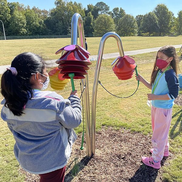 Two young girls wearing face masks playing outdoor musical instruments shaped like flowers in their school playground