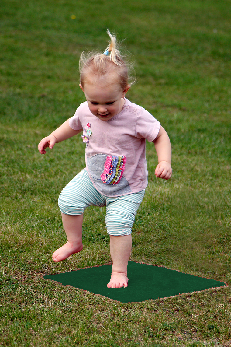 a young girl jumping on a green musical stepping stone square in a lawn which is disguising musical notes