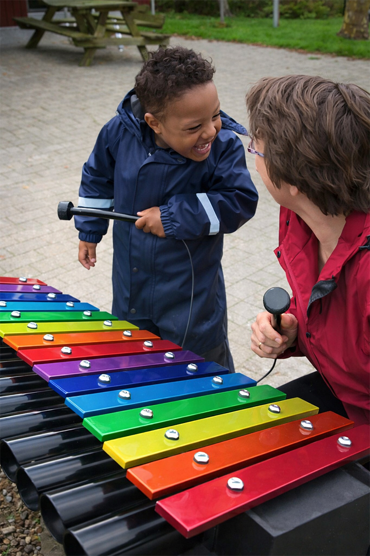 a young boy with additional needs playing a rainbow ourdoor xylophone and laughing with his teacher kneeled beside him