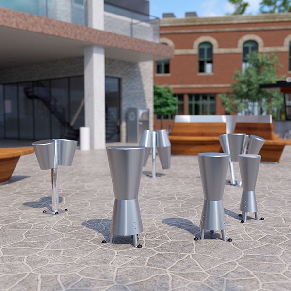 A group of modern stainless steel outdoor drums on a stone-paved plaza near a coffee shop and wooden benches, with a staircase in the background.
