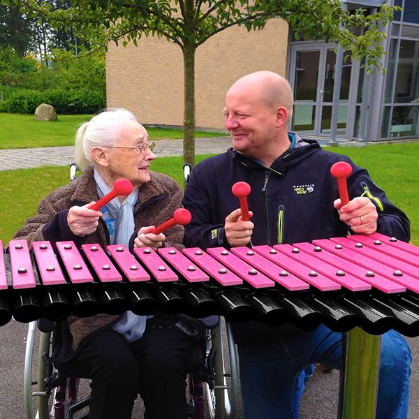 Man kneeling next to an elderly lady in a wheelchair playing an outdoor xylophone