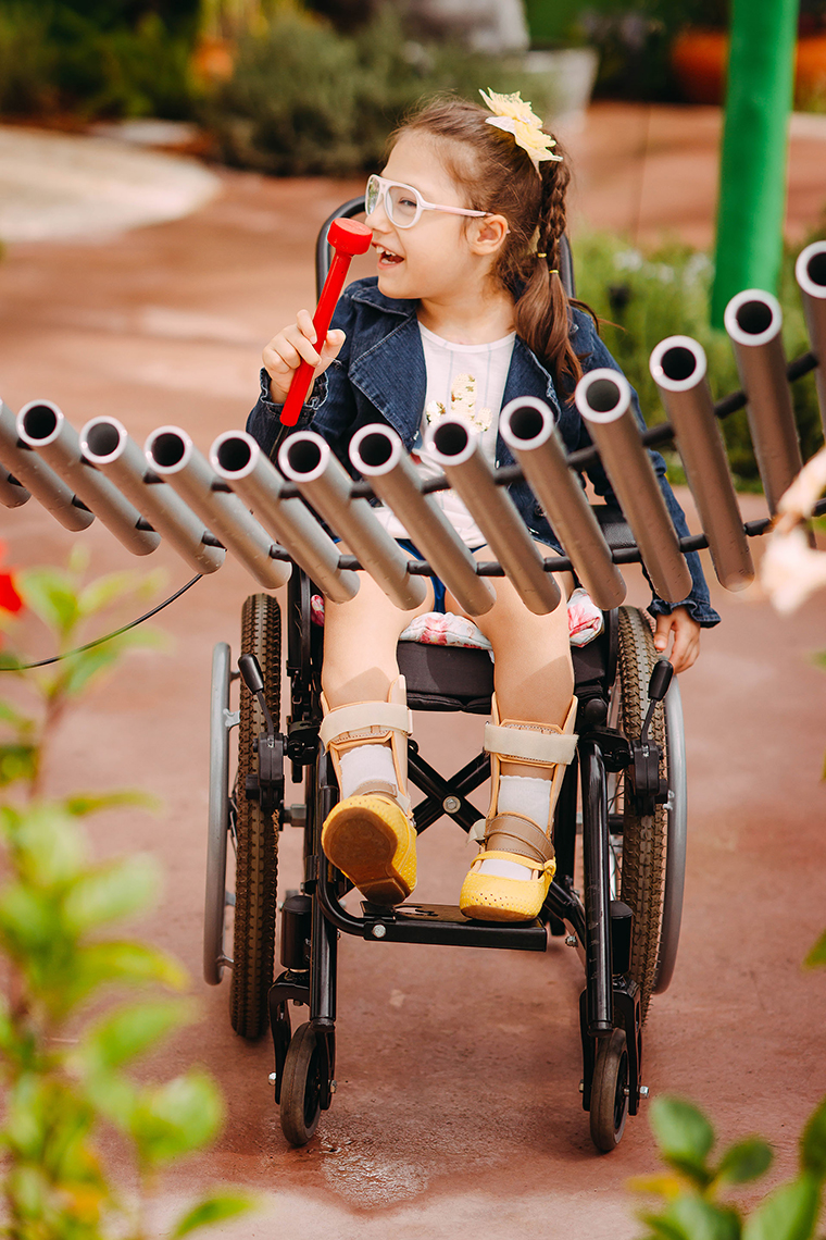 a young girl with special needs and in a wheelchair smiling and playing a large metal outdoor musical instrument in a playground