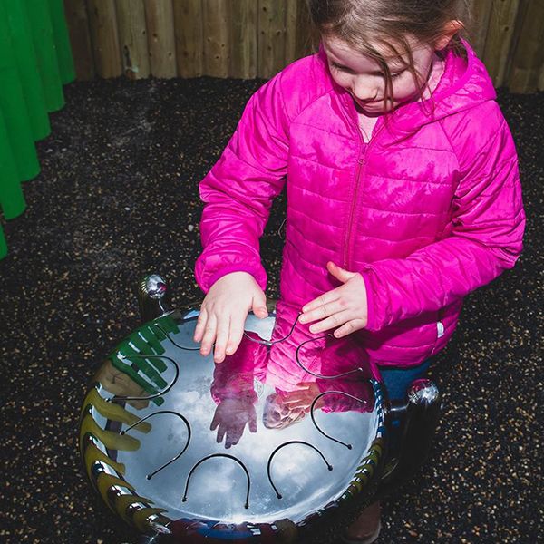 a little girl wearing a bright pink coat playing a stainless steel tongue drum in a music playgound