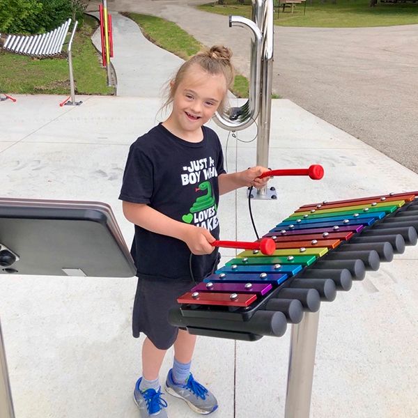 a young girl with special needs playing a rainbow colored outdoor xylophone in a music park at summer camp 