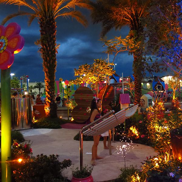 two girls playing a silver outdoor xylophone in a playground at night with LED lights in the plants and trees 