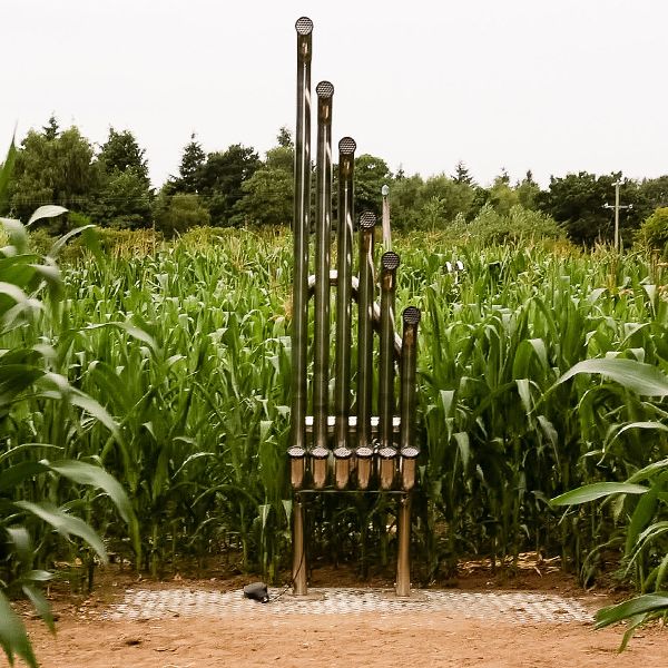 a tall outdoor musical pipe instrument called an aerophone made of six stainless steel pipes standing in a corn field