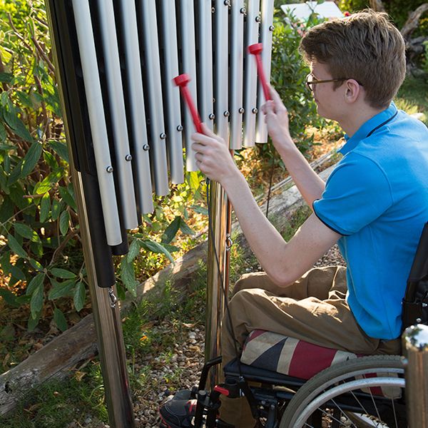 A young man in a wheel chair reaching up and playing a large outdoor musical instrument in a music garden