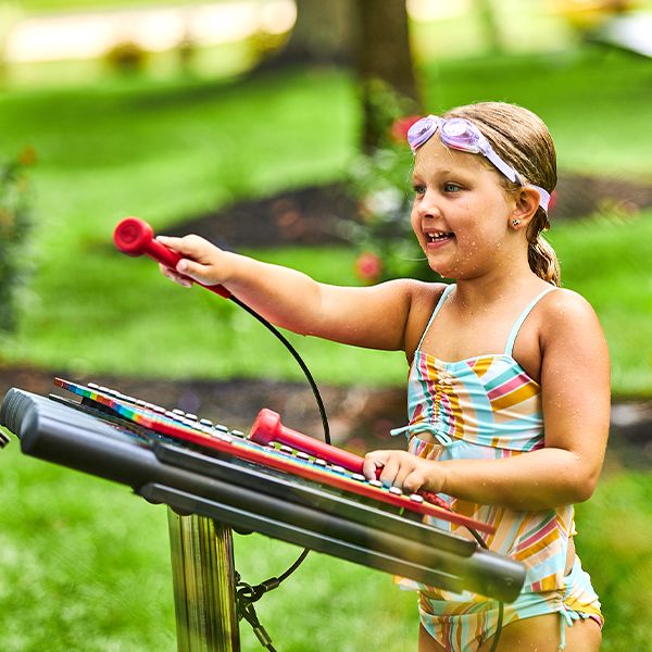 a young girl wearing a swimming costume and goggles on top of her head playing a large colourful outdoor xylophone 