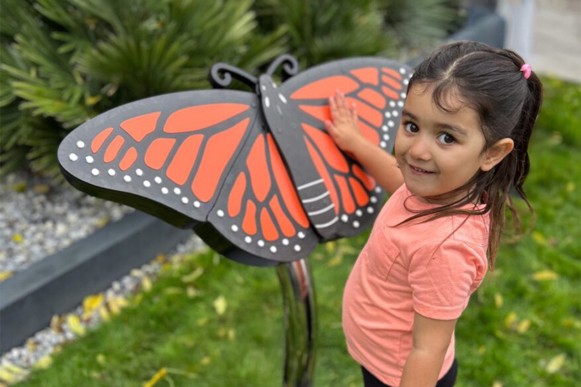 A child in a pink shirt interacts with a large, colorful monarch butterfly music sculpture in a garden setting.