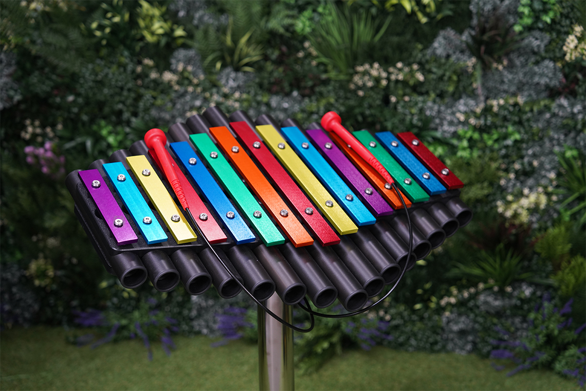 an outdoor xylophone with rainbow colored notes on a stainless steel leg with a green leafy background