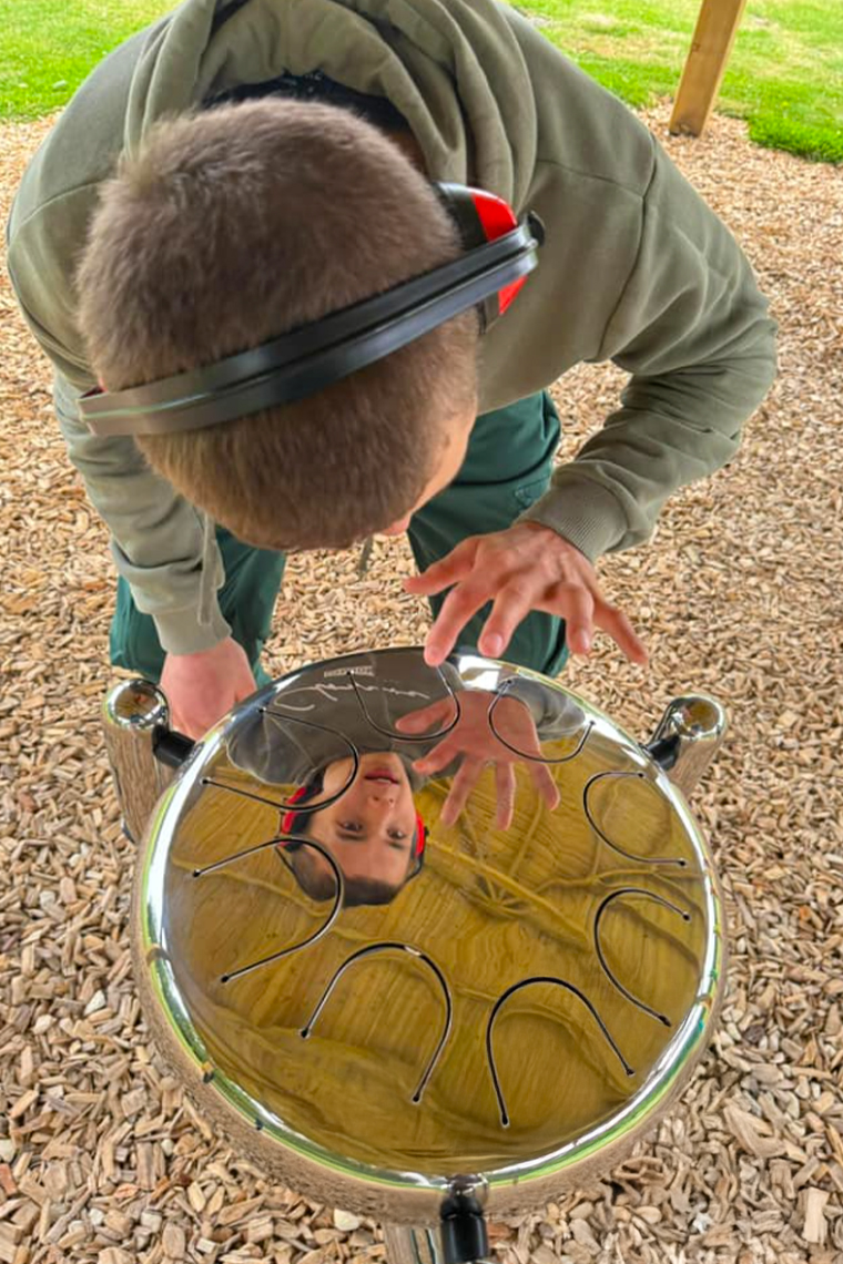 A teenage boy in a music barn wearing headphones playing a stainless steel tongue drum while looking at his reflection