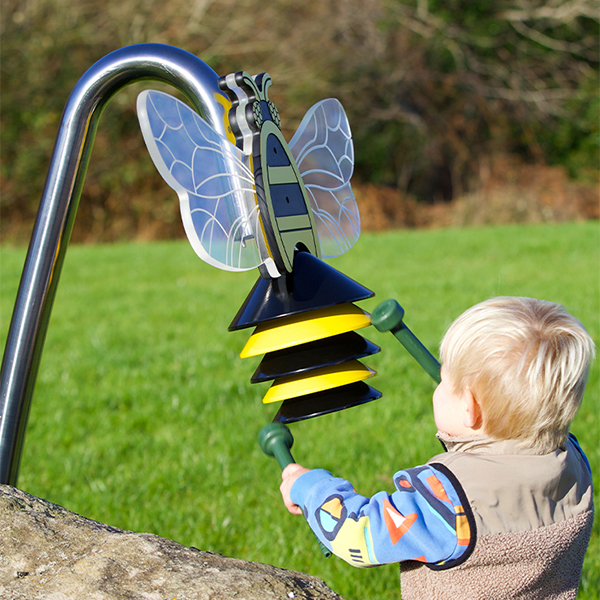 an outdoor musical instrument shaped like a bumblebee on a stainless steel post being played by a young blond boy