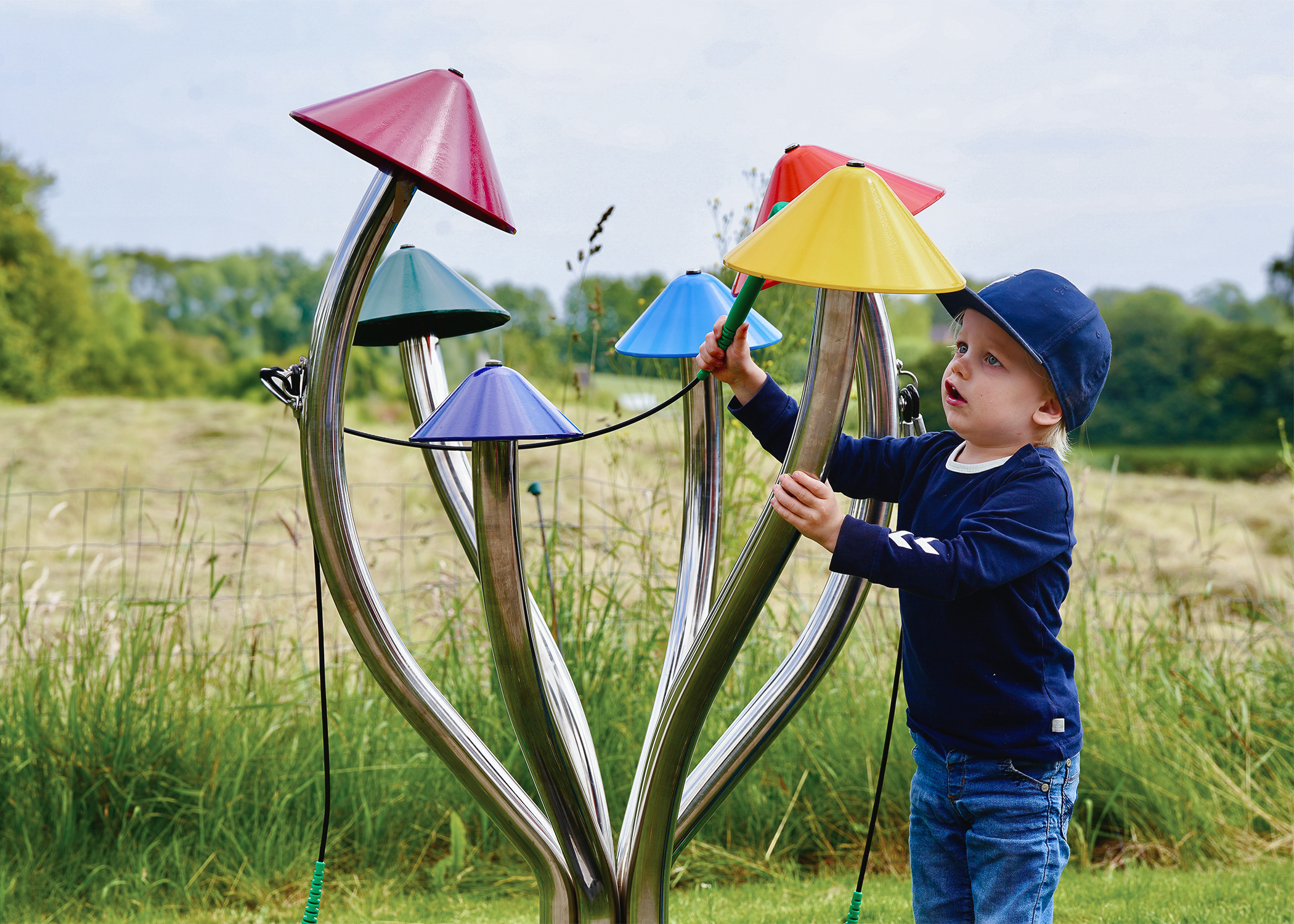 a young boy wearing a cap playing a large outdoor musical instrument shaped like field mushrooms with different coloured caps on each stalk