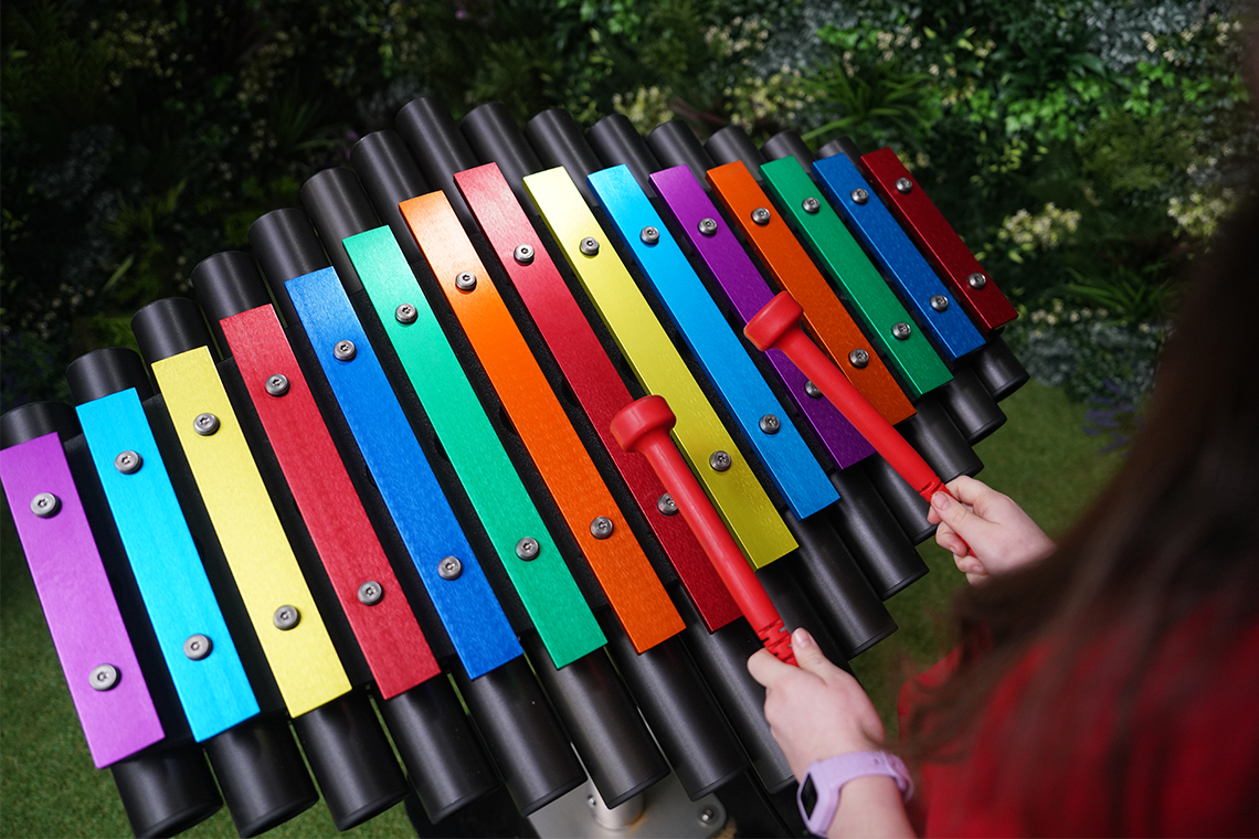 an outdoor xylophone with rainbow colored notes on a stainless steel leg with a green leafy background