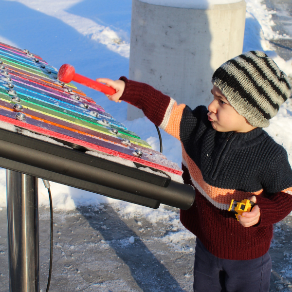 a young boy in a hat playing a rainbow colored outdoor musical xylophone outside the clinton macomb public libary