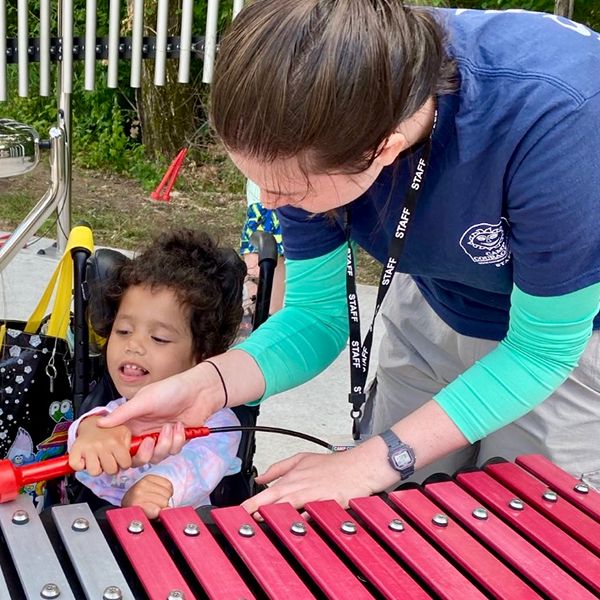 a young disabled girl in a wheelchair being helped by a young female carer to play an outdoor xylophone in a music park at summer camp