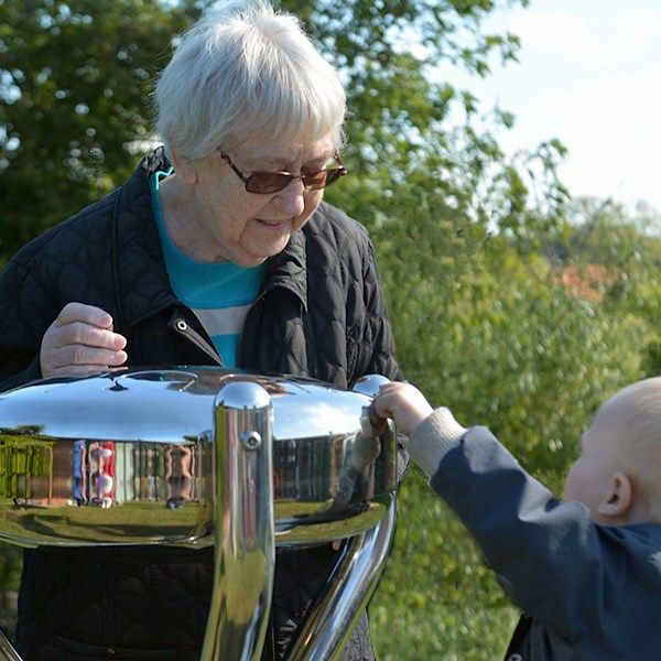 Older lady playing on an outdoor stainless steel tongue drum with hergrandchild