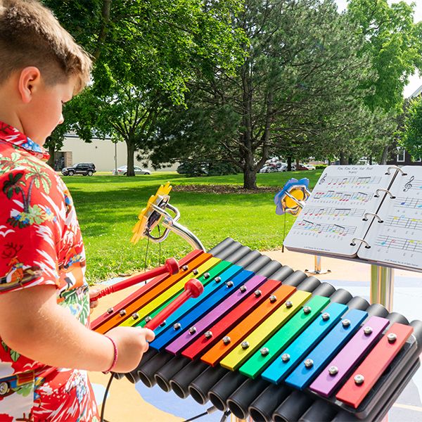 a young boy playing a large outdoor rainbow colored xylophone with a separate music book on a stand in a music park