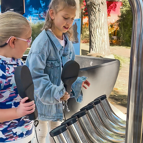 Two young girls are outdoors playing large tubular musical instruments with paddles. They appear engaged and joyful in a sunny park setting.