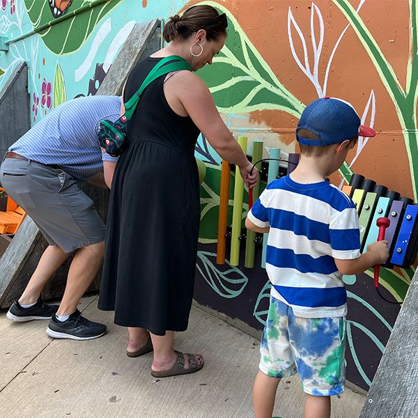 a set of three outdoor musical instruments fixed to a wall with a colorful mural being played by a family of three