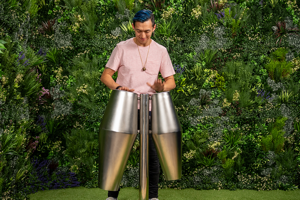 A person with blue hair plays large stainless steel percussion drum instruments outdoors, surrounded by lush green foliage, conveying a lively, vibrant atmosphere.
