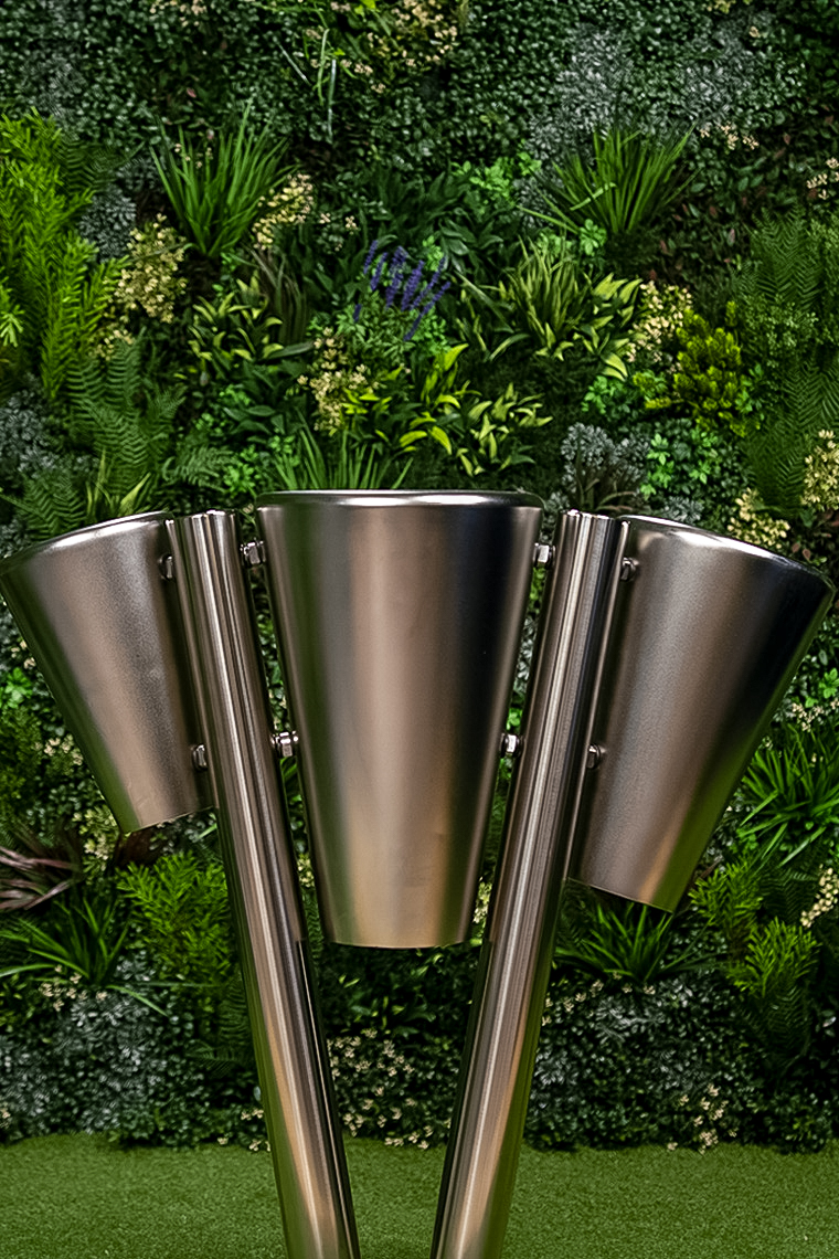 A stainless steel outdoor drum  with three cylindrical heads on a single post in a garden setting. The background features lush green foliage and plants.