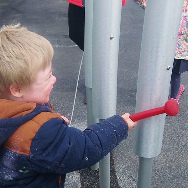 young boy hitting large outdoor tubular bells with a red beater in special needs school playground