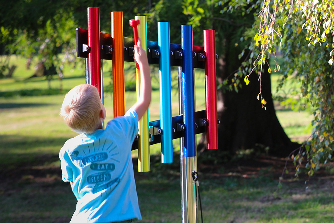 Young boy reaching up and playing a rainbow coloured set of six musical chimes in a school playground