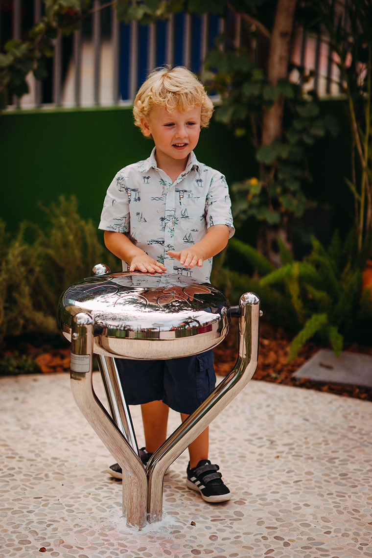 A blonde little boy playing a stainless steel tongue drum in a music park or playground