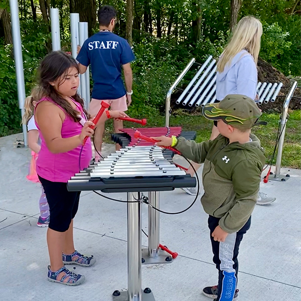 a young boy and girl standing either side of and playing a large outdoor xylophone in a music park at summer camp 