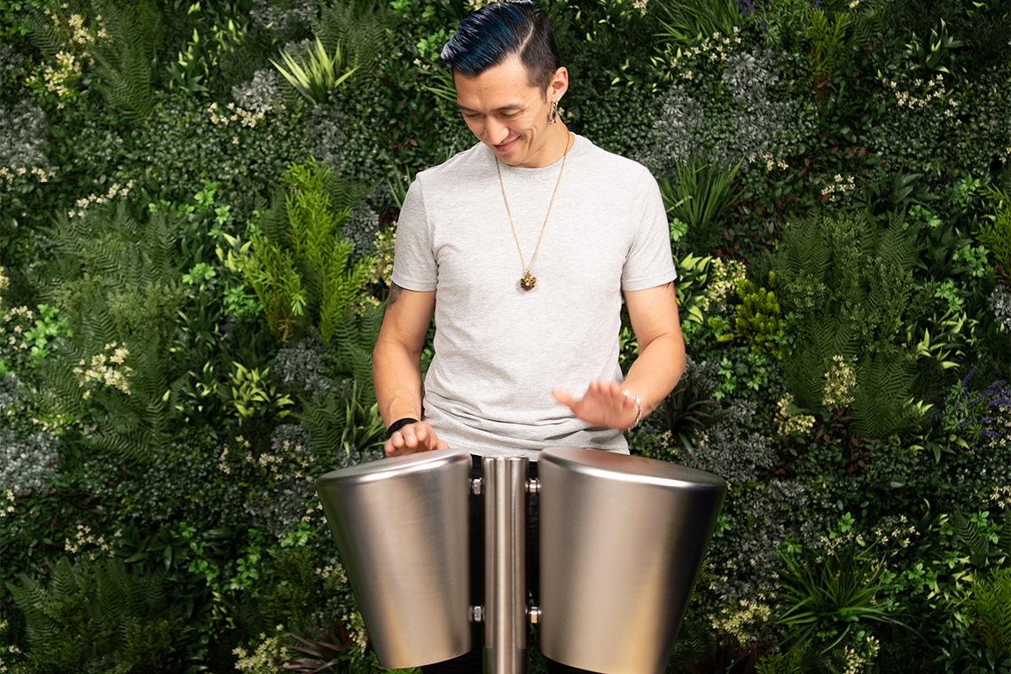 A person with blue hair plays silver steel drums joyfully against a lush, green wall of plants. The scene conveys a vibrant and lively atmosphere.