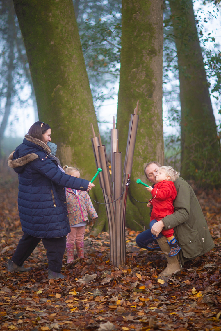 a family of four playing on some outdoor musical chimes shaped like cattails in a woodland