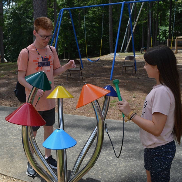 two teens one male one female playing a large colorful outdoor musical instrument shaped like field mushrooms in a camp playground