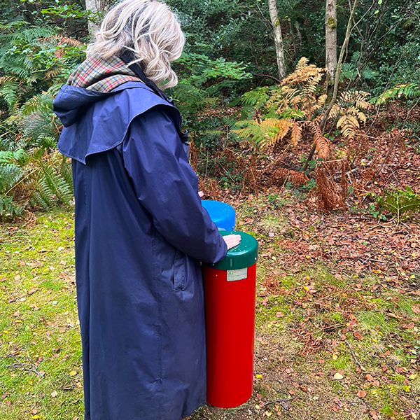 a lady in a long blue coat standing and playing a pair of outdoor conga drums in a woodland setting