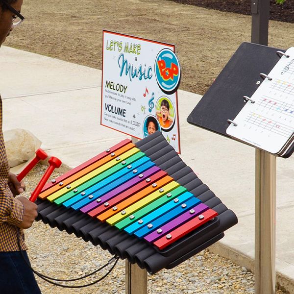 a large outdoor xylophone with rainbow colored notes along with an outdoor music book next to a sign saying lets make music