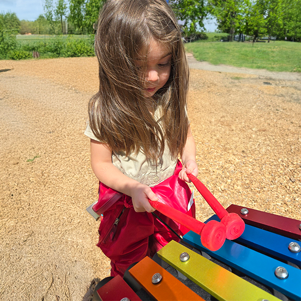 A young girl playing a small outdoor metallophone in rainbow colors in a school playground