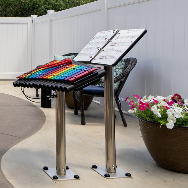 an outdoor xylophone with rainbwo colored notes along with an outdoor music book on a stainless steel leg in a s senior living courtyard