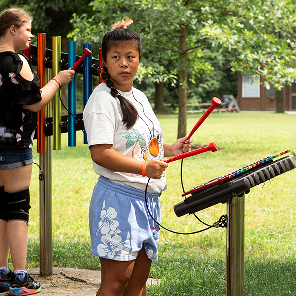 a teenage girl playing an outdoor xylophone in a percussion park at summer camp with other instruments a players in the background
