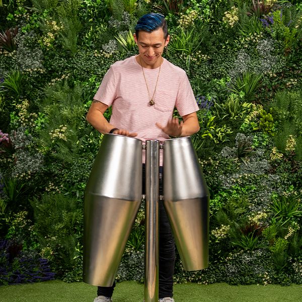 A person with blue hair plays large, metal percussion drum instruments outdoors, surrounded by lush green foliage, conveying a lively, vibrant atmosphere.