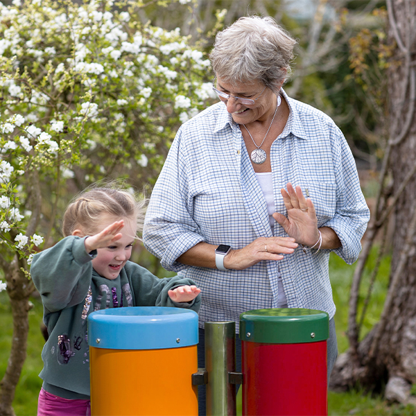 A grandmother and granddaughter playing an outdoor musical drum with trees in blossom behind
