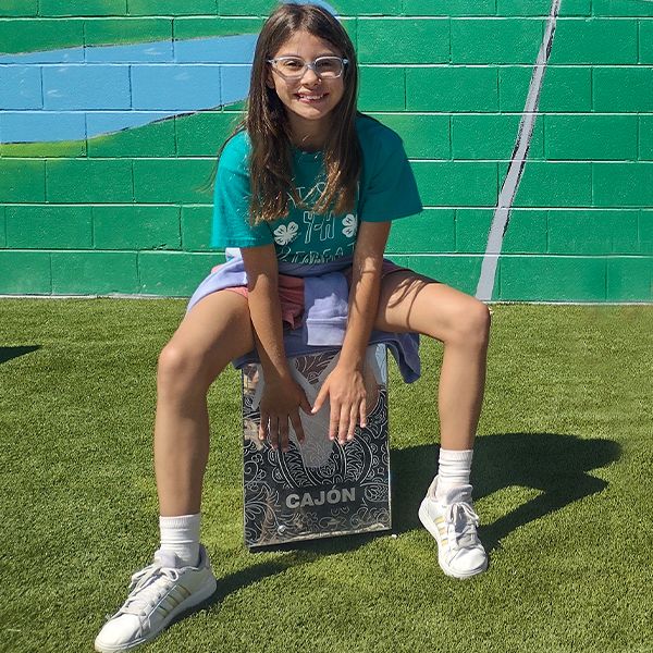 a teenage girl sitting on and playing a silver outdoor cajon drum at the town of versailles outdoor music garden