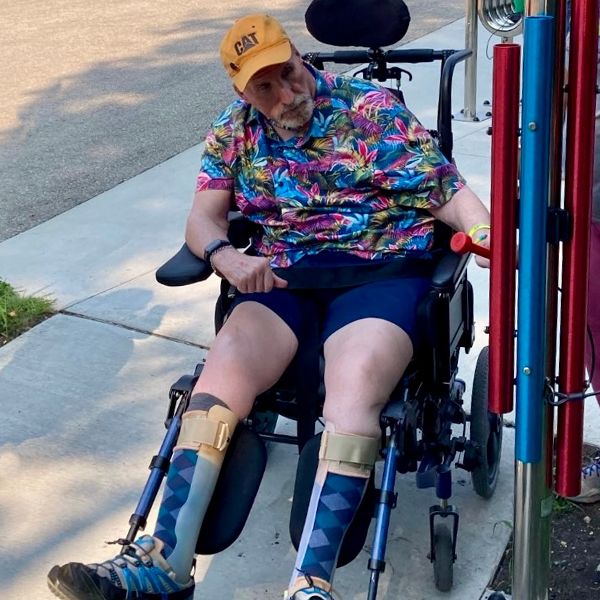 older man in a wheelchair and with legs in braces playing a large set of rainbow chimes in a music park at summer camp