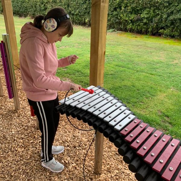 A girl wearing headphones playing an outdoor xylophone with pink and silver notes at the Thomas Centre music pavilion.