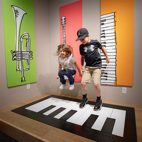 a boy and a girl in caps jumping on a large floor piano in the NAMM museum