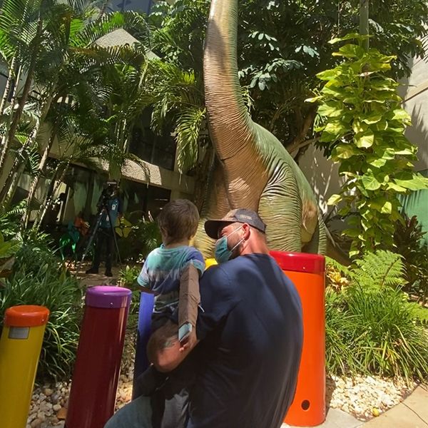 a father kneeling down and holding his son in front of five colored outdoor drums and a dinosaur sculpture